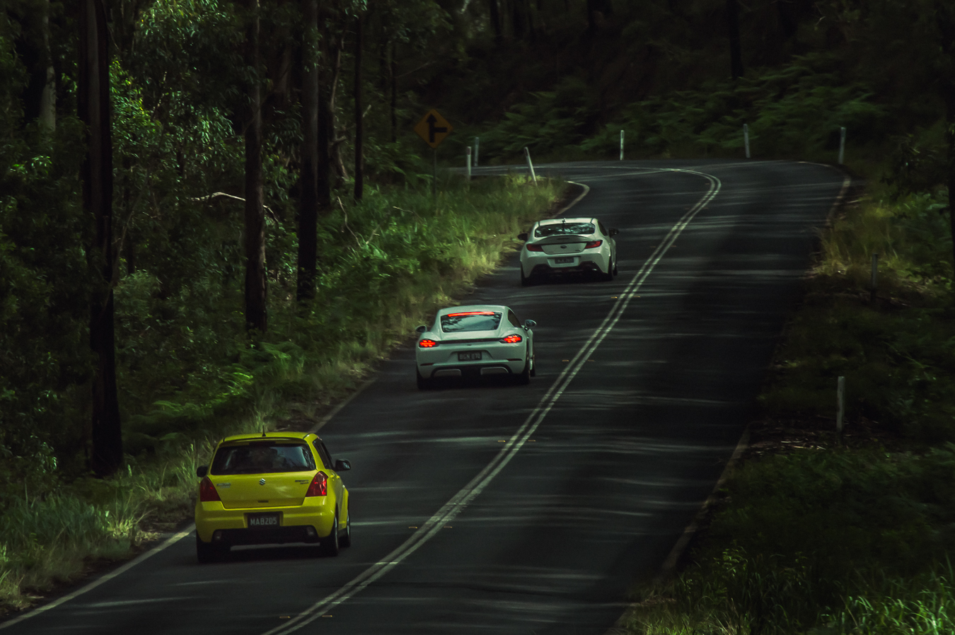 A yellow hatchback and sports cars on a tree lined road